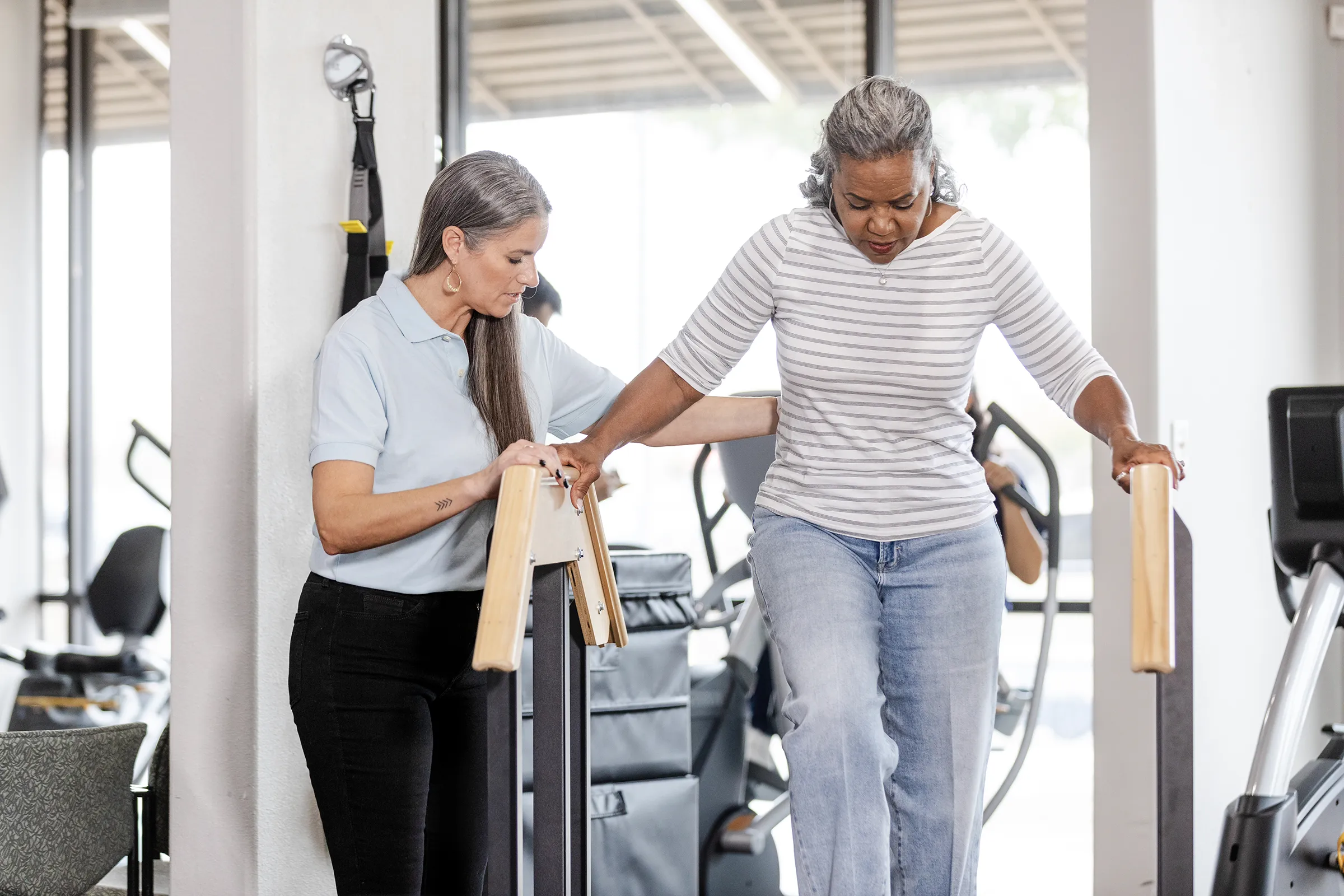 Trainer assists woman using parallel bars in rehabilitation gym